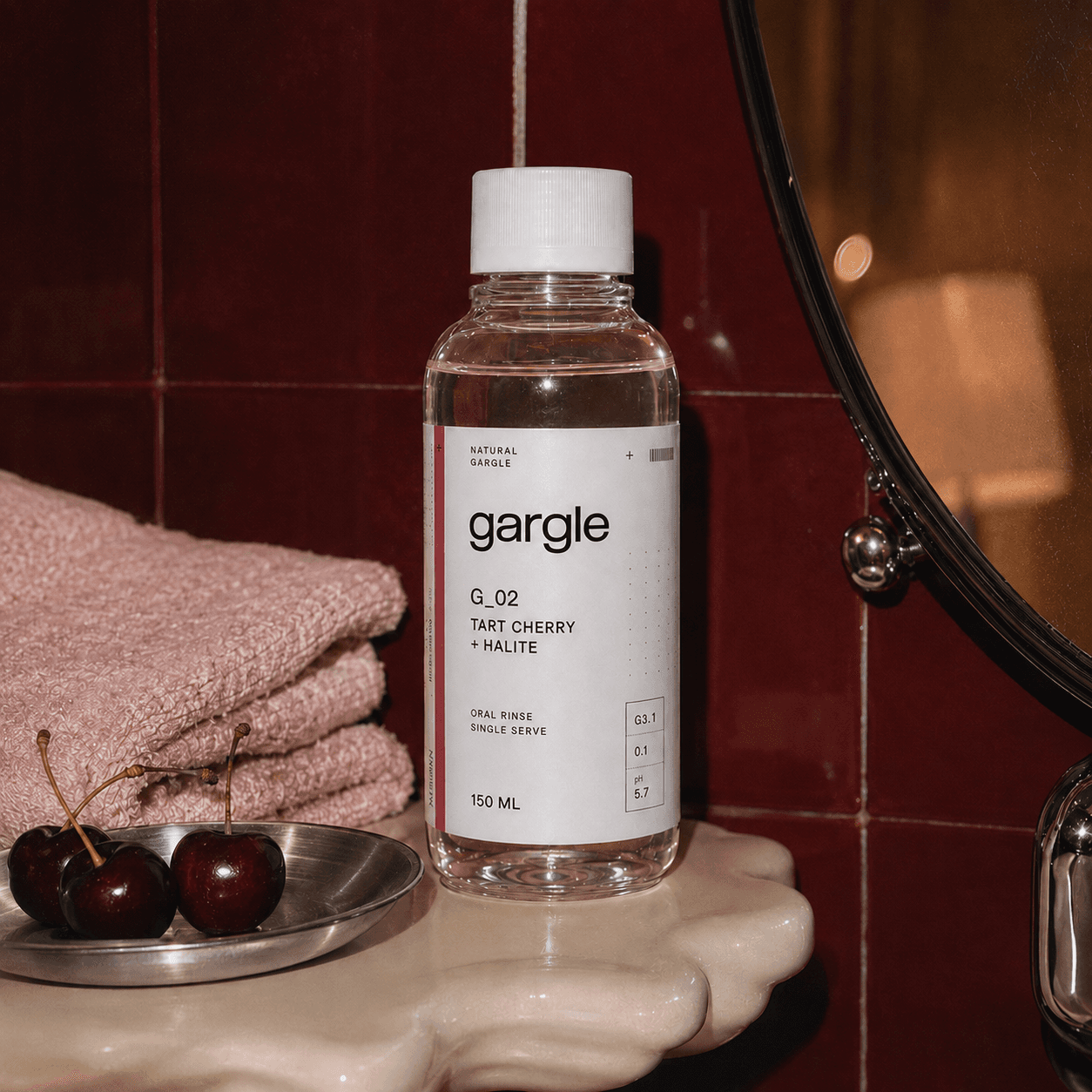 Gargle Tart Cherry + Halite bottle beside fresh cherries on a burgundy-tiled vanity.